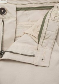 Pantalons beiges avec une fermeture éclair et un bouton, dotés de poches zippées doubles et d'accents en tissu vert contrastant à l'intérieur.