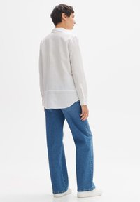 Camicia bianca in lino a maniche lunghe con colletto a punta, abbinata a jeans in denim larghi di colore blu chiaro e sneakers bianche.