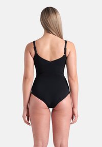 Vrouw met lang blond haar in een zwarte badpak gezien vanaf de achterkant tegen een effen lichte achtergrond.