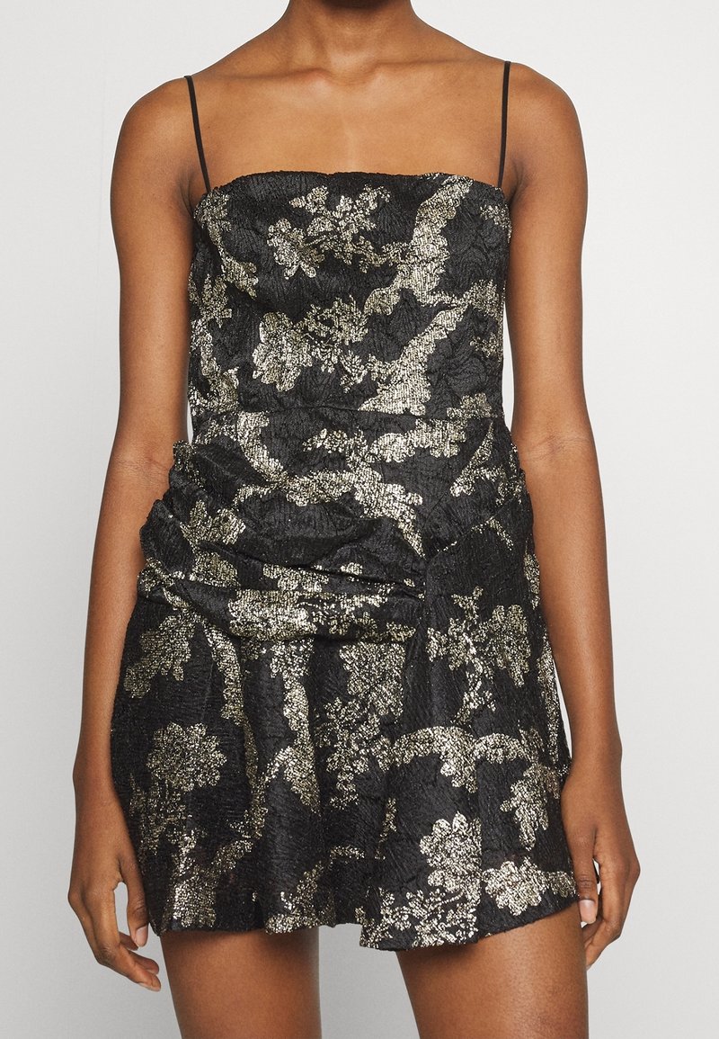 Robe noire avec un motif floral en métal doré, tissu texturé, corsage ajusté et jupe à volants. Dotée de fines bretelles spaghetti.