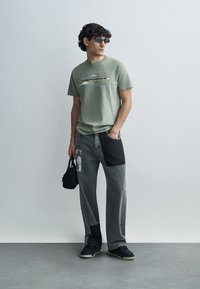 Πράσινη κοντομάνικη μπλούζα με γραφιστικό σχέδιο, σε συνδυασμό με γκρι τζιν παντελόνια που διαθέτουν κομμάτια υφάσματος και πάνελ αντίθετου χρώματος. Μαύρα αθλητικά παπούτσια και τσάντα.