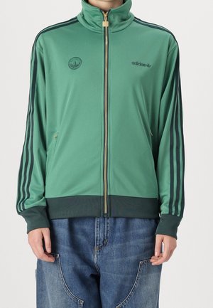 Person iført en grøn Adidas trackjakke med sorte striber på ærmerne, lynet op, kombineret med blå jeans.