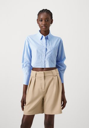 Cropped blue striped shirt with button closure and long sleeves paired with beige pleated shorts featuring a belt detail.