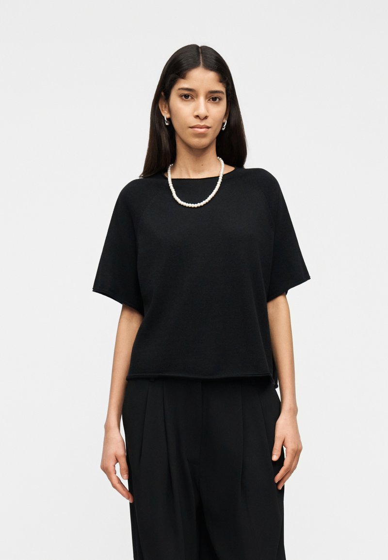 Femme aux longs cheveux noirs portant un haut noir à manches courtes, un pantalon plissé noir, un collier de perles et des boucles d'oreilles argentées en forme de créoles, debout devant un fond blanc.