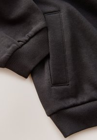 Gros plan sur une manche de sweat-shirt noire avec un poignet côtelé et une poche latérale fendue sur un fond neutre.