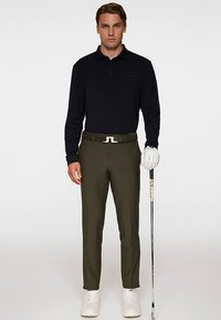 Czarna koszulka polo z długim rękawem, oliwkowe spodnie, biały pasek z logo, białe tenisówki, trzymający kij golfowy. Prosty fason i dopasowany styl.