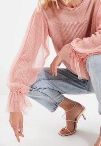 Bleknad rosa, strukturerad stickad tröja med genomskinliga volangärmar, matchat med ljusblå, korta jeans och strapazierade beige högklackade sandaler.