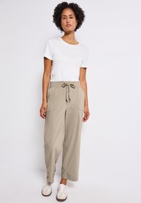 Losse beige broek van zachte stof, met een elastische tailleband en trekkoord, en twee voorkeurszakken. Gecombineerd met een wit T-shirt.