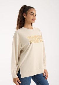 Jasno beżowy sweatshirt o luźnym kroju, z długimi rękawami, okrągłym dekoltem i błyszczącym graficznym wzorem z przodu. Miękka tekstura materiału.