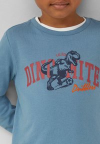Blaues Langarm-Sweatshirt mit einem Dinosaurier-Graphic, dem Text "Dino-mite" und sportlichen Akzenten in Rot und Orange. Weicher Stoff mit geripptem Ausschnitt.