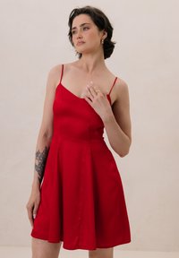 Czerwona sukienka w kształcie litery A z cienkimi ramiączkami, wykonana z gładkiego satynu, z dopasowanym gorsetem i rozkloszowanym dołem. Zauważalny detal bocznego szwu.