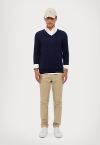 Suéter de cuello en V navy sobre una camisa blanca con cuello, pantalones khaki claros y zapatillas blancas. Gorra beige con logo en la parte frontal.