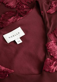 Lähivõte burgundi värvi kangast, millel on tekstuuriga lilleline broderie ja valge riideetikett, millel on kiri "NOELLA" ning suurus väike.