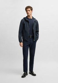 Marinblå lättvikts zip-up jacka med huva, bärs över en matchande marinblå t-shirt och slim-fit byxor, ihop med svarta snörskor.