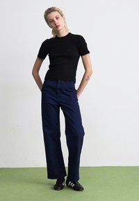 Personne debout, les mains dans les poches, portant une chemise noire à manches courtes, un pantalon large marine et des baskets noires avec des rayures blanches.