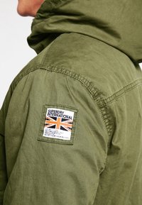 Veste verte avec un patch cousu sur le haut du bras représentant un drapeau Union Jack et le texte "Superdry International".
