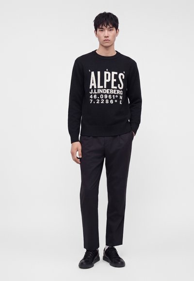 Jeune homme debout portant un pull noir avec le texte Alpes en blanc et des coordonnées, pantalon noir et baskets noires sur un fond uni.