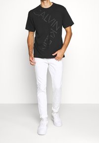 Musta puuvillainen t-paita, jossa on harmaa vinottain oleva "CALVIN KLEIN" -logo, yhdistettynä valkoisiin kapealahkeisiin farkkuihin ja valkoisiin lenkkareihin, yksiväristä taustaa vasten.