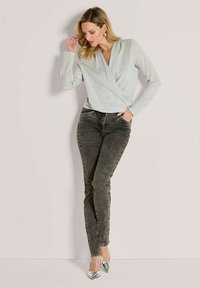 Hellgrünes, langärmliges Wickeloberteil mit einem subtilen Glanz, kombiniert mit dunkelgrauen Skinny-Jeans und silbernen spitzen Pumps.