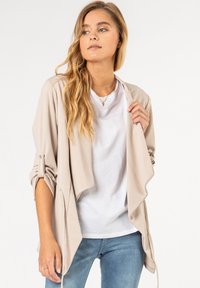 Beige satijnen jack met een gedrapeerde voorkant, opgerolde mouwen en strikdetail. Gedragen over een eenvoudige witte blouse en blauwe jeans.