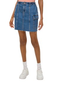 Jupe en denim avec une taille élastique, deux poches avant, un ourlet effiloché et des coutures verticales, de couleur bleu moyen. Associée à des baskets blanches.