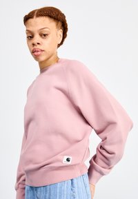 Sweatshirt rose clair avec un col rond, des poignets et un ourlet côtelés. Présente un petit patch logo Carhartt sur le côté gauche. Texture douce.