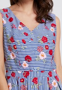 Vestido sin mangas en rayas azules y blancas con bordado floral en rojo, rosa y morado. Escote en V y cintura fruncida con pliegues.