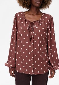 Brązowa bluzka w kropki z wiązaniem przy szyi, długimi bufkami i guzikami z przodu. Gładki materiał, luźny krój i kieszonka na piersi.