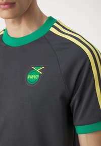 Mörkgrå fotbollströja med gröna och gula detaljer. Har en räfflad krage och det jamaicanska fotbollsfederationens logga på bröstet.