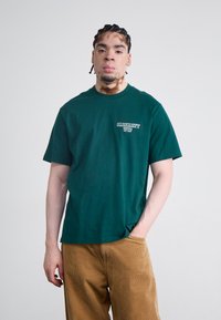 Grønn bomull t-skjorte med rund hals, med en liten hvit "Carhartt WIP" logo på venstre side, kombinert med lysebrune cordbukser.
