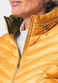Veste matelassée jaune avec un col haut, dotée d'une fermeture éclair, d'accents en matériel doré et d'une doublure verte contrastante.