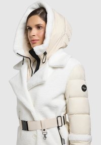 Bílá bunda s plyšovou texturou, s tlustou, zateplenou béžovou kapucí, páskem v pase a detailním logem na rukávu.