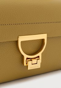 Clutch em couro verde-oliva com acabamento texturizado, apresentando um fecho circular em tom dourado e um detalhe subtil de costura.