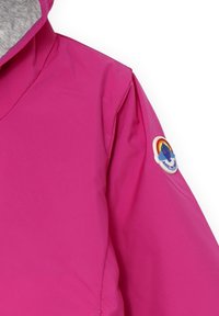 Giacca impermeabile rosa con design a V. Presenta un interno grigio e una colorata toppa circolare sulla manica con un motivo di arcobaleno e montagna.