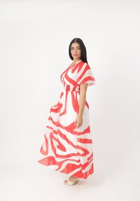 Vestito maxi in tessuto bianco con audaci motivi ondulati rossi. Maniche corte, vita aderente e gonna fluida. Abbinato a sandali con tacco.
