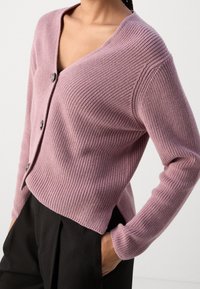 Jasnofioletowy kardigan zapinany na guziki z prążkowanej tkaniny, z dekoltem w serek i długimi rękawami; dopasowany w talii z bocznymi rozcięciami. Widać czarne spodnie.