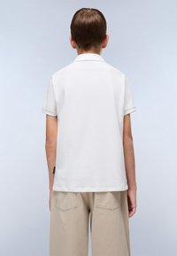 Hvit polo shirt med korte ermer, krage og rett fald, kombinert med beige, løstsittende bukser. Merket er synlig på side-sømmene.