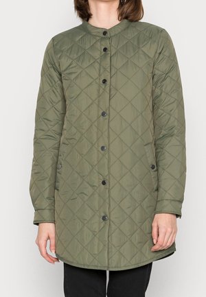 Mujer con chaqueta larga acolchada de color verde oliva con botones a presión negros y dos bolsillos laterales, de pie frente a un fondo liso.