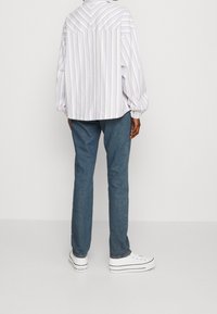 Camicia a righe in viola chiaro e bianco, vestibilità comoda con colletto, abbinata a pantaloni in denim blu e sneakers bianche.
