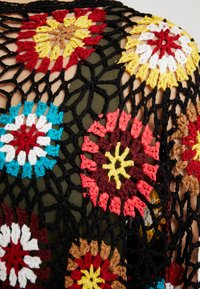 Haut en crochet avec motifs floraux colorés en rouge, jaune, bleu et blanc, avec un design en filet noir. Le tissu sous-jacent est vert olive.