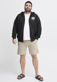 Svart hettejakke med glidelås og frontlogo, hvit T-skjorte under, beige shorts og brune sandaler. Stoffet ser mykt og uformelt ut.