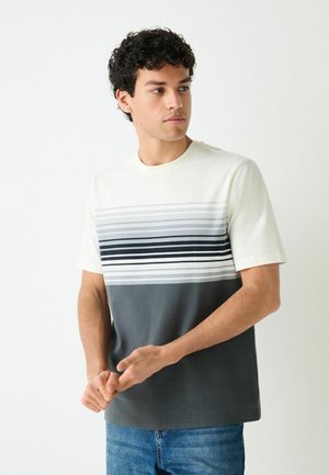 Young man with curly hair wearing a short-sleeve shirt with horizontal gray and black stripes, looking to the side against a white background.