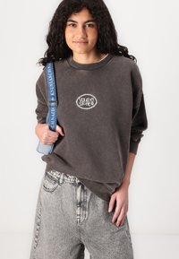 Dark grey oversized sweatshirt with a white logo at the center, paired with light grey patterned jeans and a blue strap bag.