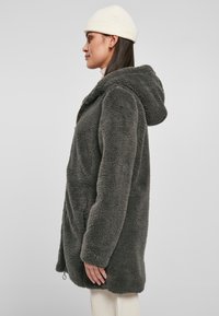 Sivi plišani fleece jakna s kapuljačom, s patentnim zatvaračem sprijeda, bočnim džepovima i mekanom teksturom; nosi se preko svijetlih hlača.