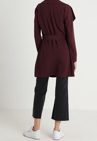 Bordowy płaszcz zapinany w talii, z długimi rękawami i drapowanym tyłem. Połączony z czarnymi, krótkimi dżinsami i białymi trampkami.