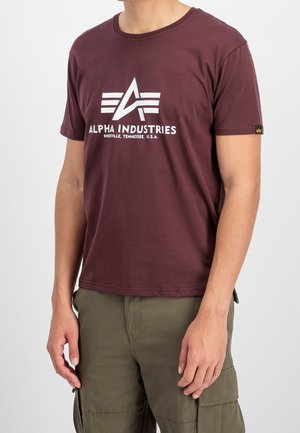 Mann trägt ein mahagonifarbenes Alpha Industries T-Shirt mit Logo und Schriftzug, kombiniert mit olivgrünen Cargohosen, steht vor einem schlichten Hintergrund.