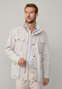 Giacca beige chiaro realizzata in materiale leggero, dotata di zip e bottoni a presión, due ampie tasche sul petto e colletto alto.