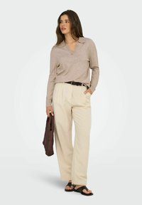 Mujer vestida con una blusa de manga larga beige con cuello y pantalones anchos crema, sosteniendo un bolso marrón acolchado, de pie con una mano en el bolsillo.