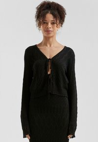 Schwarze gestrickte Strickjacke mit einem kurzen Design, das einen tiefen V-Ausschnitt und einen Bindeschluss aufweist. Strukturiertes Diamantmuster und lange Ärmel.