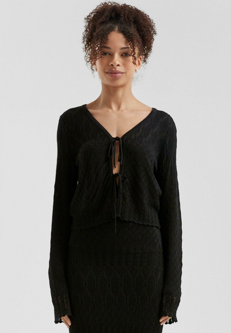 Schwarze gestrickte Strickjacke mit einem kurzen Design, das einen tiefen V-Ausschnitt und einen Bindeschluss aufweist. Strukturiertes Diamantmuster und lange Ärmel.